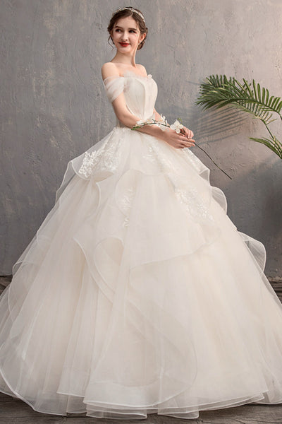 A woman wearing a voluminous off-the-shoulder white tulle wedding gown with floral lace appliques and a layered ruffled skirt, standing against a textured gray wall with a green plant in the background.